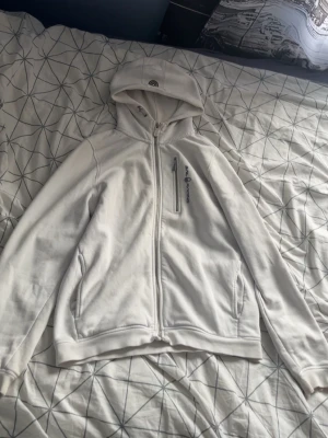 Vit hoodie från Sail Racing - Säljer en vit hoodie från Sail Racing med dragkedja framtill och en bröstficka med dragkedja. Hoodien har broderad logga på bröstet och en klassisk huva. Tillverkad i mjukt material, perfekt för chill dagar. Håll i vänstra ärmen men det märks knappast och lite färg som inte heller märks