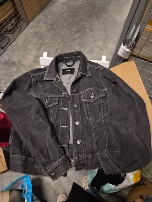 Svart jeansjacka från Bik Bok - Svart jeansjacka från Bik Bok med klassisk design, knappar framtill och två bröstfickor med lock. Jackan har snygga sömdetaljer och är tillverkad i slitstarkt jeansmaterial. Storlek s