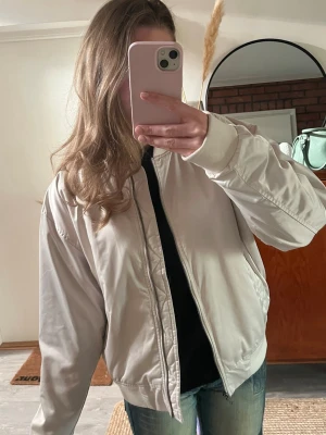 Vit bomberjacka med dragkedja - Snygg vit bomberjacka med klassisk passform och dragkedja framtill. Jackan har ribbade muddar vid ärmslut och midja samt två sidofickor. Perfekt för dig som vill ha en clean och stilren look. Materialet är glansigt och ger en modern känsla.