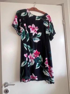 Svart blommig kortklänning med knyt - Svart kortklänning med stora rosa och vita blommor samt gröna blad. Klänningen har korta ärmar och ett knytband i midjan för en snygg passform. Perfekt för sommarens alla tillfällen och har en mjuk, lätt känsla.