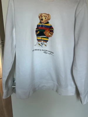 Vit hoodie Polo Bear Ralph Lauren - Vit hoodie från Ralph Lauren med ikoniskt Polo Bear-motiv på framsidan. Björnen har färgglada kläder och håller en basketboll. Hoodien har dragsko i huvan och ribbade muddar. Perfekt för dig som gillar streetstyle och klassiska märken.