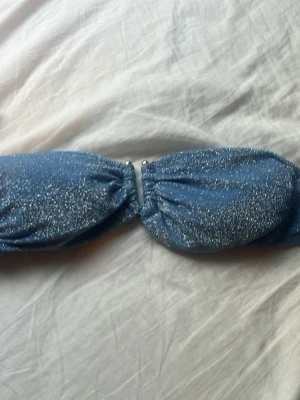 Blå glittrig bandeau bikini - Säljer en blå bandeau bikini med glittereffekt. Överdelen har rynkade detaljer och metallringar i mitten, medan underdelen är klassisk med samma glittriga material. Perfekt för dig som vill sticka ut på stranden!