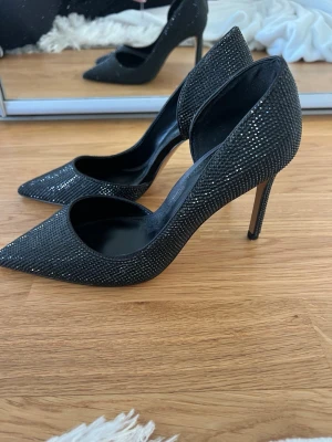 Svarta pumps med strass från Mango - Helt oanvända!! Snygga svarta pumps från Mango med spetsig tå och höga smala klackar. Skorna är täckta med små glittrande strassdetaljer som ger en lyxig känsla. Materialet är textil med syntetisk insida. Perfekta för att höja vilken outfit som helst. Klackhöjd ca 10cm