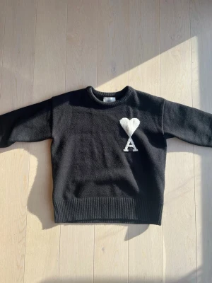 Svart stickad tröja från Ami Paris - Svart stickad tröja från Ami Paris med rund halsringning och ribbade muddar. På bröstet finns ett vitt hjärta och bokstaven 'A' i kontrastfärg. Perfekt för dig som gillar stilrena och trendiga plagg.