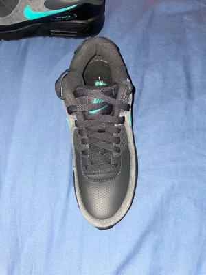 Nike Air Max 90 svart/grå med turkos swoosh - Nike Air Max 90 sneakers i svart och grått med turkosa detaljer och klassisk turkos swoosh på sidan. Ovandel i en mix av läder och textil, robust sula med synlig Air Max-enhet. Perfekt för dig som gillar streetwear och sportig stil.