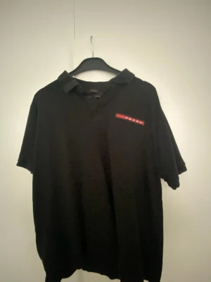 Svart kortärmad skjorta från Prada - Säljer en svart kortärmad skjorta från Prada med klassisk krage och knappslå framtill. På bröstet sitter en ikonisk röd logotyp med PRADA-tryck. Skjortan har en loose passform och är perfekt för en stilren look. Den har krympt lite.