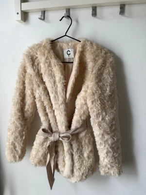 Krämvit fuskpälsjacka med knytskärp - Säljer en fluffig krämvit fuskpälsjacka med snyggt knytskärp i midjan. Jackan har lång ärm, mjukt foder och en klassisk öppen krage. 