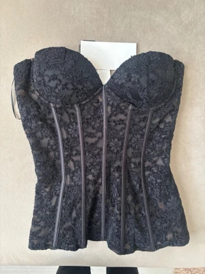 Svart spetskorsett från Zara - Supersnygg svart korsett från Zara med vadderade kupor och dekorativ spets över hela plagget. Korsetten har markerade sömmar framtill för en extra formande effekt och transparenta partier i spetsen. Perfekt att styla till olika outfits för en trendig look.