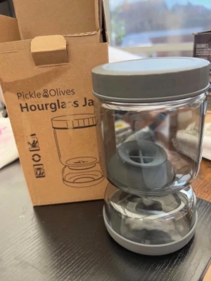 Pickle & Olives Hourglass Jar - Säljer en Pickle & Olives Hourglass Jar i genomskinlig plast med grå lock och insats. Perfekt för att förvara oliver eller pickles, smart design med två fack och enkel hällfunktion. Levereras i originalkartong.
