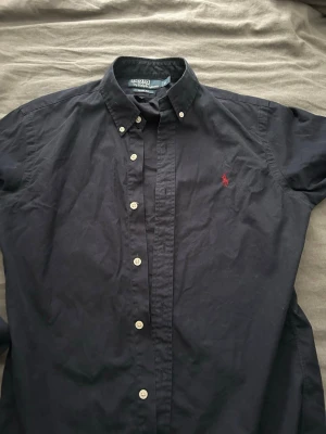 Mörkblå skjorta från Ralph Lauren - Klassisk svart skjorta från Ralph Lauren med button-down krage och vita knappar. Skjortan har lång ärm och den ikoniska röda logotypen broderad på bröstet. Denna skjorta passar perfekt på festliga aktiviteter och är ett måste i garderoben! 
