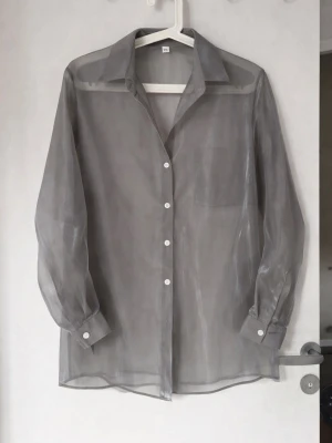 Transparent skjorta i organza / sheer oanvänt ny - Transparent skjorta i organza / sheer modell i grå nyans. Lätt och luftig med lös passform och knappar framtill. Perfekt över linne, topp eller bikini. Trendig och elegant. Mycket fint skick.