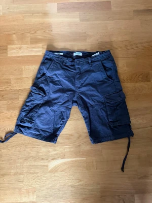 Blå cargoshorts med snörning -  par blå cargoshorts med flera fickor och snörning. Shortsen är loose och är perfekta för sommaren. Tillverkade i bomull för en skön känsla. Perfekta för dig som gillar en casual stil.