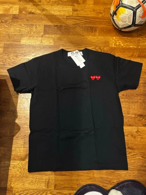 Svart Comme des Garçons PLAY t-shirt - Svart t-shirt från Comme des Garçons PLAY med de ikoniska röda hjärt-applikationerna på bröstet. Klassisk rund hals och korta ärmar. Tillverkad i mjuk bomull för skön känsla och stilren look.