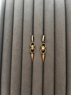 Guldtonade örhängen med konform - Säljer ett par trendiga örhängen i guldtonad metall med två ringar, en liten glittrande detalj och en konformad hängande spets. Örhängena har en modern och edgy design som sticker ut. Perfekta för dig som gillar unika accessoarer.