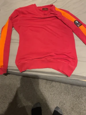 Röd stickad tröja från Parajumpers - Man. Snygg röd stickad tröja från Parajumpers med orange och gula ränder längs ärmarna. Tröjan har rund halsringning, ribbade muddar och en patch på ena ärmen. Perfekt för dig som vill sticka ut med färg och stil.