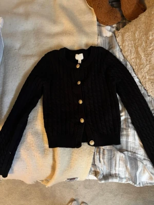 Svart kabelstickad kofta från H&M - Snygg svart kabelstickad kofta från H&M med rund halsringning och guldknappar framtill💕