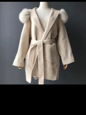 Feminina of sweden coat  - Stilren beige kappa med huva och fluffig äkta päls runt axlarna. Kappan har knytskärp i midjan, stora fickor och rak passform. Perfekt för dig som vill ha en trendig och mysig look under kalla dagar.