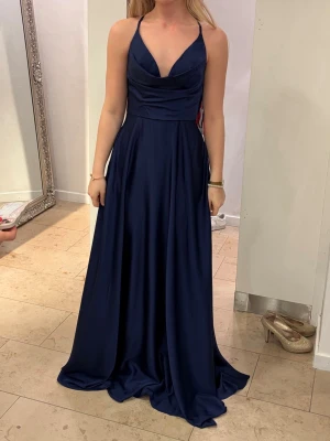 Marinblå långklänning med öppen rygg - Elegant marinblå långklänning med djup v-ringning och tunna axelband som korsas i ryggen. Klänningen har en markerad midja och ett flytande, svepande fall. Perfekt för bal eller festliga tillfällen. Materialet har en lätt glansig finish.