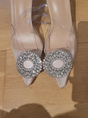 Klara pumps med strass och transparent detalj - Säljer ett par snygga pumps med spetsig tå och transparent plastmaterial. Skorna har en dekorativ cirkel av glittrande strass framtill och en smal klack. Perfekta för dig som vill sticka ut med en trendig och elegant look.
