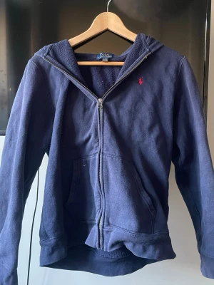Mörkblå zip up hoodie från Ralph Lauren - Mörkblå hoodie från Ralph Lauren med dragkedja framtill och två fickor. Klassisk design med liten röd logga på bröstet. Tillverkad i mjukt material som känns skönt mot huden. XL i unisex barn storlek så passar s/m. Varan är lite nopprig och har ett litet hål på höger ärm❣️
