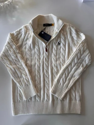 Vit kabelstickad tröja Polo Ralph Lauren - Kabelstickad vit tröja från Polo Ralph Lauren med halv dragkedja och hög krage. Tröjan har ribbade muddar och nederkant samt den klassiska broderade loggan på bröstet. Perfekt för lager-på-lager och snygg till jeans.