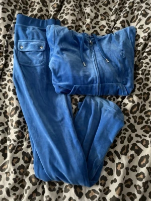 Blå velour loungewear-set Juicy Couture - Säljer ett blått velourset från Juicy Couture med zip-hoodie och matchande byxor. Hoodien har dragsko och broderad logga på bröstet, byxorna har fickor med knappar bak. Perfekt för chill dagar hemma eller när du vill vara extra comfy.