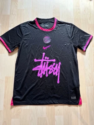 Svart PSG x Stüssy t-shirt Nike - Säljer en svart PSG x Stüssy t-shirt från Nike med rosa detaljer och tryck. T-shirten har korta ärmar, rund hals och stora loggor framtill. Perfekt för dig som gillar streetwear och sportiga vibes.
