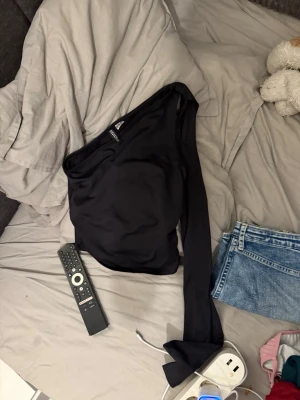 Svart  topp från HM - Snygg svart  topp från Divided med lång ärm på ena sidan och axelbandslös på andra. Tillverkad i mjukt stretchigt material som sitter tight och markerar siluetten. Perfekt för dig som vill ha en trendig och unik look.