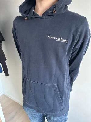 Mörkblå hoodie från Scotch & Soda - Mörkblå hoodie från Scotch & Soda med vit logga på bröstet. Hoodien är helt plain och tillverkad i mjuk bomull, perfekt för en avslappnad stil. 