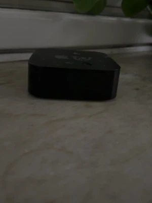 Apple TV med fjärrkontroll - Säljer en svart Apple TV med tillhörande fjärrkontroll. Boxen har en stilren, fyrkantig design i plast och fjärren är smal med touchyta och tydliga knappar för enkel navigering. Perfekt för streaming och appar direkt till din TV.
