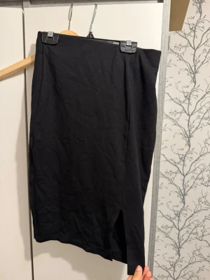 Svart kjol med slits från Amisu - Säljer en svart kjol från Amisu i storlek 36. Kjolen har en enkel och stilren design med en snygg slits framtill som ger en trendig touch. Perfekt att matcha med en crop top eller stickad tröja för en cool look.