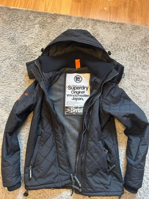 Svart vindjacka från Superdry - Snygg svart vindjacka från Superdry med quiltat mönster, huva och dragkedja framtill. Jackan har orange detaljer, två sidofickor med dragkedja och en mjuk grå insida. Perfekt för blåsiga dagar och streetwear-stil.