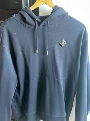 Svart hoodie från J.Lindeberg - Snygg svart hoodie från J.Lindeberg med huva och dragsko. Klassisk känguruficka framtill och diskret logomärke på bröstet. Tillverkad i mjukt material som känns skönt mot huden. Perfekt för en avslappnad och stilren look.