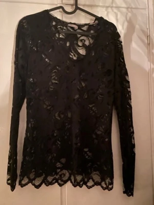 Svart spetsblus med lång ärm - Säljer en svart blus i genombruten spets med lång ärm och rund hals. Blusen har fina spetsdetaljer och vågig kant nertill, perfekt för dig som gillar en elegant och trendig look.