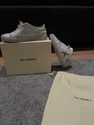 Vita sneakers Axel Arigato fågelbrodyr - Snygga vita sneakers från Axel Arigato med färgglad fågelbrodyr på sidan, storlek 40-41. Klassisk låg modell med snörning och stilren design. Skorna är tillverkade i skinn och kommer med originalbox samt dustbag. Skorna har lite skador men inga stora. Skorna är perfekt nu till våren samt sommaren. Skriv om funderingar. Pris ej hugget i sten! ÄKTA! TVÄTTAR SÅKLART INNAN JAG SKICKAT DET!