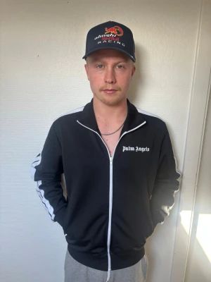 Palm Angels svart zip-tröja - Palm Angels svart zip-tröja med vit dragkedja och logga på bröstet. Tröjan har vita detaljer längs axlar och ärmar, samt ribbade muddar. Perfekt för en sportig och avslappnad look.
