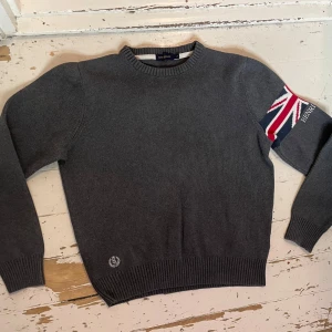 Grå stickad tröja Henri Lloyd S - Snygg grå stickad tröja från Henri Lloyd i storlek S. Tröjan har lång ärm och coolt Union Jack-mönster i rött, vitt och blått på ena ärmen med broderad logga. Ribbad mudd vid hals, ärmslut och nederkant. Den är knappt använd men har ett litet hål i ena armhålan (se bild)