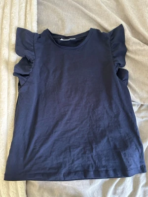Mörkblå topp med volangärmar - Snygg mörkblå topp med volangdetaljer vid ärmarna. Rund halsringning och rak passform. Perfekt för dig som vill ha en enkel men ändå trendig look. Tillverkad i mjuk bomull som känns skön mot huden.