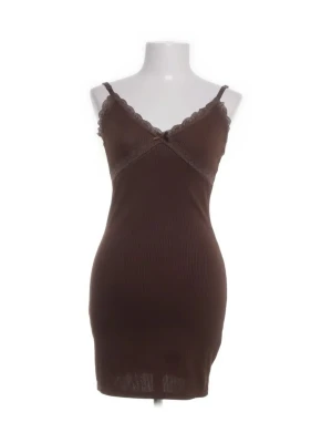 Brun ribbad spetsklänning - Söt brun kortklänning med smala axelband och spetsdetaljer vid urringningen. Klänningen är ribbad och har en liten rosett framtill för extra detalj. Figurnära passform och ärmlös design, perfekt för dig som gillar en enkel men ändå snygg look.