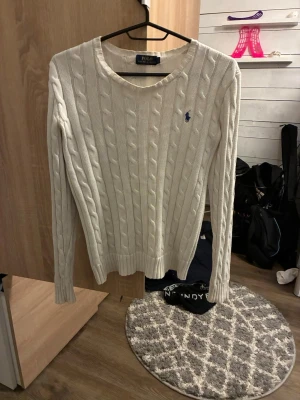 Vit kabelstickad tröja Polo Ralph Lauren - Snygg vit kabelstickad tröja från Polo Ralph Lauren med klassisk rund halsringning och blå broderad logga på bröstet. Tröjan har långa ärmar och ribbade muddar vid ärmslut och nederkant. Perfekt för dig som gillar stilrena och tidlösa plagg.