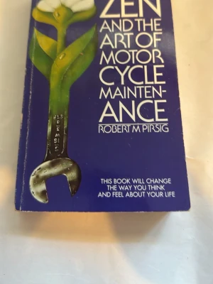 Zen and the Art of Motorcycle Maintenance - WEn klassisk roman av Robert M. Pirsig med blått omslag och vit text. Boken har ett unikt omslagsmotiv med en blomma och en skiftnyckel. Perfekt för dig som gillar filosofiska berättelser och motorcyklar.