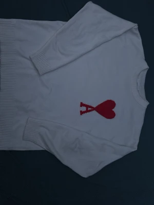 Vit Ami Paris tröja  - Säljer en vit stickad tröja med rund hals och ribbade muddar. På bröstet finns ett rött hjärta och bokstaven 'A' i samma färg. Tröjan har en clean look och är perfekt för dig som gillar stilrena detaljer.
