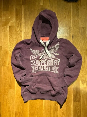  Superdry med tryck - Snygg  hoodie från Superdry med stort vitt tryck framtill med texten 'Superdry Real Athl.'                  Storlek S  
