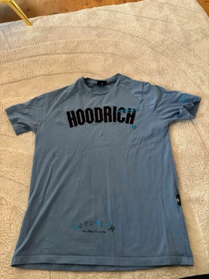 Blå HOODRICH t-shirt i bomull - Snygg blå t-shirt från HOODRICH med svart tryck framtill och textdetaljer på ryggen. Tillverkad i mjuk bomull och har en klassisk passform med korta ärmar och rund hals. Perfekt för en avslappnad streetwear-look.