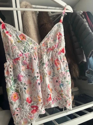 Blommig spetslinnetopp med broderi - Söt blommig linnetopp med tunna axelband och v-ringning. Toppen är vit i grunden med färgglada broderade blommor och spetsdetaljer. Perfekt för sommaren och ger en romantisk vibe till din outfit.