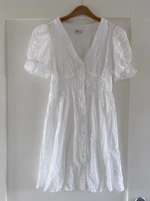 Vit kort klänning med puffärm - Söt vit kortklänning med broderade detaljer och puffärmar. Klänningen har v-ringning, knappar framtill och är tillverkad i luftig bomull. Perfekt för sommarens alla tillfällen och ger en romantisk vibe med sina volanger och spetsliknande struktur.