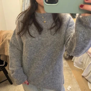 Grå oversized stickad tröja - Mysig grå stickad tröja med oversized passform och långa ärmar. Tröjan har en enkel design utan synliga mönster och är perfekt för lager-på-lager-stil. Materialet ser ut att vara mjukt och fluffigt, vilket ger en avslappnad vibe.