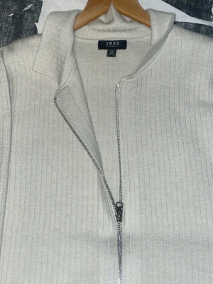 Beige ribbad ziptröja från SMOG - Stilren beige ziptröja från SMOG med ribbad struktur och hög krage. Tröjan har långa ärmar, hel dragkedja framtill och breda muddar vid ärmslut och nederkant. Perfekt för lager på lager och enkel att matcha med olika outfits.