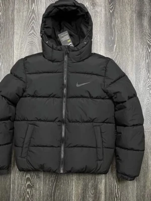 Svart pufferjacka från Nike - Säljer en svart pufferjacka från Nike med huva och dragkedja framtill. Jackan har två sidofickor och den klassiska Nike-loggan på bröstet. Perfekt för kalla dagar och riktigt skön att bära tack vare det vadderade materialet.