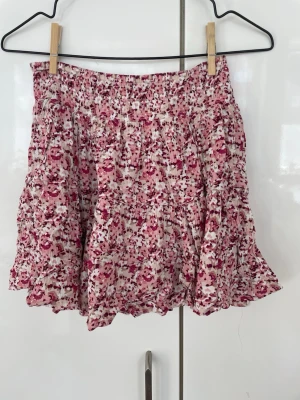 Blommig kortkjol från new yorker - Supersöt kortkjol från new yorker i vitt och rosa med blommigt mönster. Kjolen har volangdetaljer och resår i midjan för en bekväm passform samt shorts under. Perfekt för dig som gillar en feminin och lekfull stil.